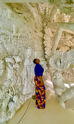 Woman looking at the inside of a white temple in Thailand. She is wearing a royal blue shirt and tropical red and yellow pants.