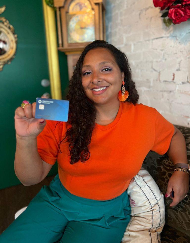 Woman wearing teal and orange. She is smiling as she is holding a blue credit card.