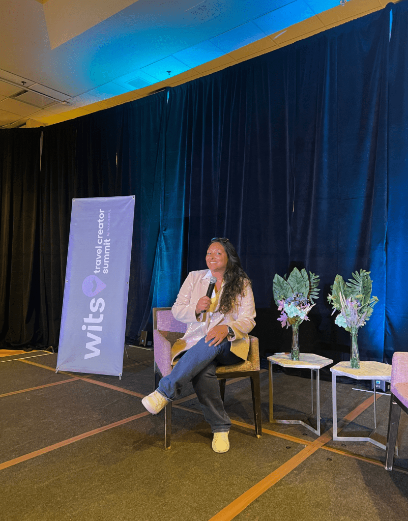 Latina Woman sitting on conference stage at WITS (women in travel summit) discussing travel conferences to attend.