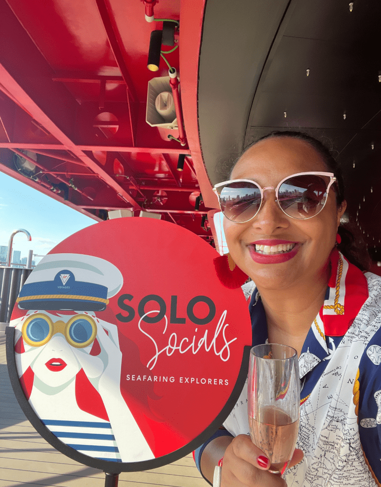 Latina Solo female traveler in a nautical-inspired outfit standing by Virgin Voyages 'Solo Socials' sign, holding a glass of champagne and ready to connect with other solo voyagers.