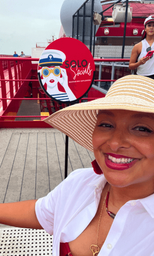 Latina Solo female traveler in a white and red outfit standing by Virgin Voyages 'Solo Socials' sign, holding a glass of champagne and ready to connect with other solo voyagers.
