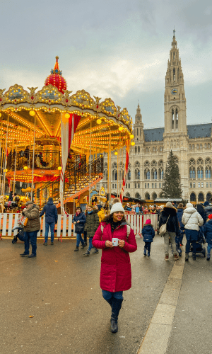 The author, a Latina Solo Female Traveler standing in front of Wiener Christkindlmarkt in Vienna, its main Christmas Market. A carousel and city hall are behind her. Why You Should Experience a European Christmas Market