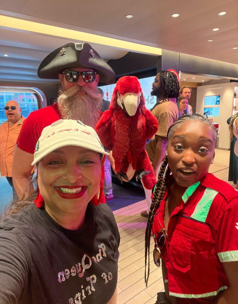 The author, wearing a black t-shirt that says the Queen of Trips posing with the Virgin Voyages Happenings Cast. A man in the background is holding a red stuffed parrot toy.