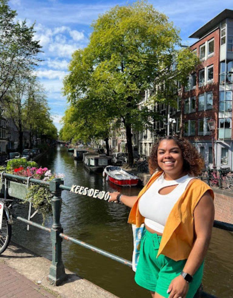 joyful Latina traveler in a mustard cardigan and green shorts poses along Amsterdam's picturesque canal, surrounded by traditional Dutch architecture and colorful flowers